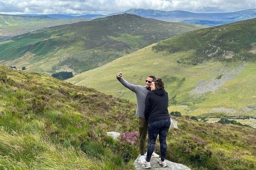 Two people taking a selfie on a grassy hill with a mountainous landscape in the background. 2