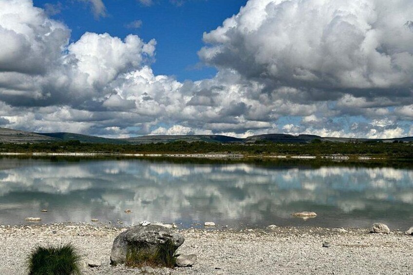A calm lake with a clear reflection of the sky and clouds, surrounded by a rocky shoreline and distant hills. 3