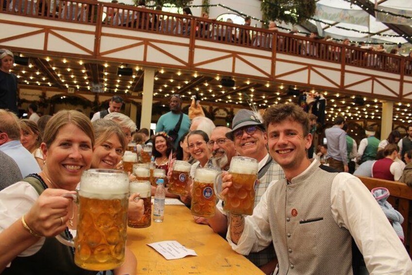 A group of people toasting with large beer mugs at a wooden table in an outdoor setting with string lights. 2