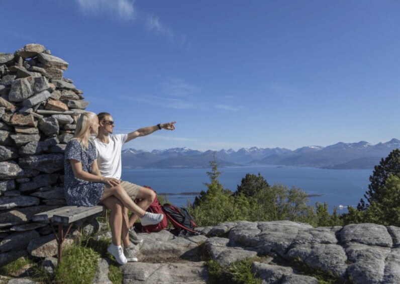 Two people sitting on a bench with a stone wall behind them, overlooking a lake and mountains. 3