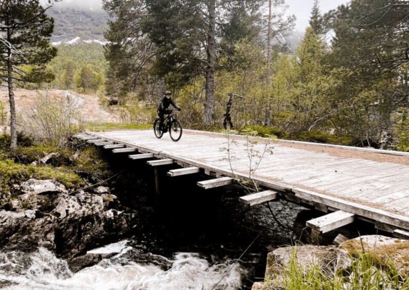 A wooden bridge over a fast-flowing river, surrounded by trees and a mountainous backdrop. 2
