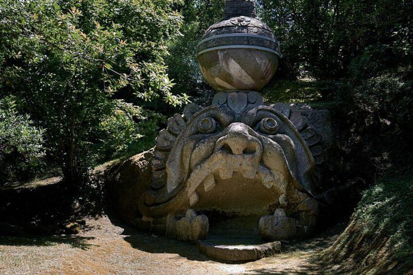 A stone sculpture of a mythical creature with a large head and a pot on top, surrounded by trees. 5
