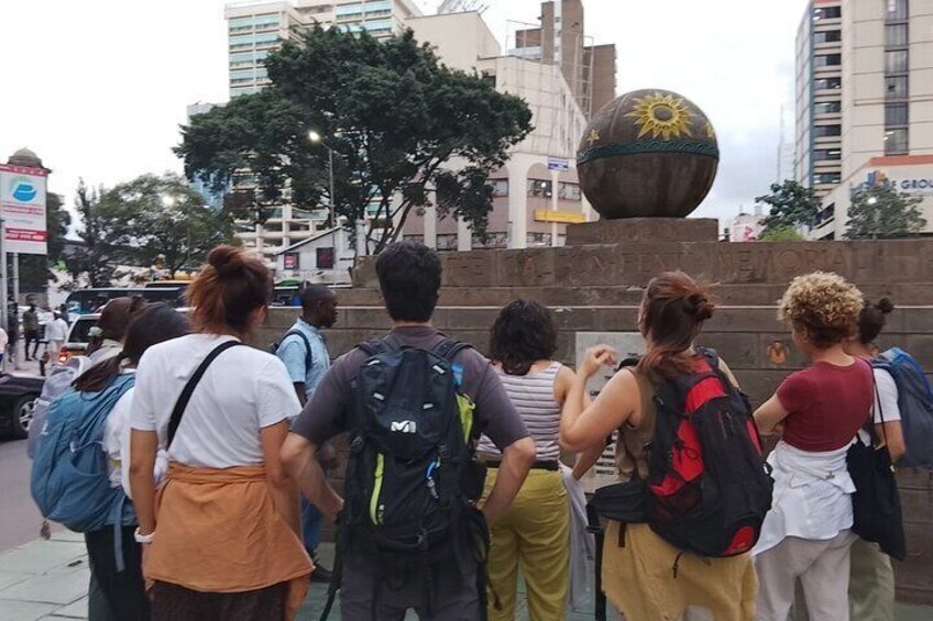 Un gruppo di persone in fila, probabilmente in attesa, con una grande scultura sferica e un edificio sullo sfondo. 4