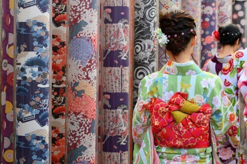 Een persoon in een kimono met bloemenmotieven, staand voor een rij traditionele Japanse textielen. 2