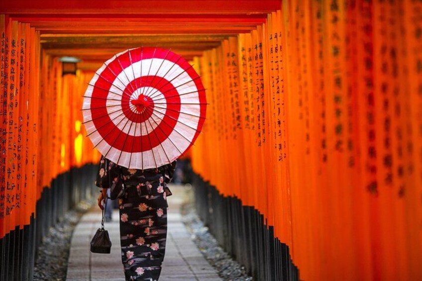 En person går ned ad en korridor kantet med orange torii-porte, mens vedkommende holder en rød og hvid paraply. 1