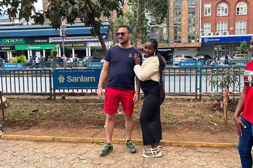 A man and a woman standing on a patterned pavement with a fence and commercial buildings in the background. 2