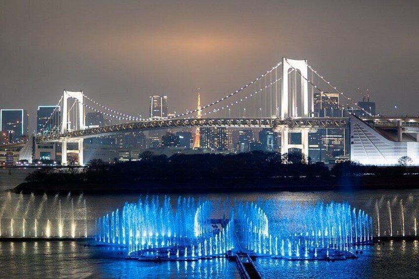 Un puente con luces iluminadas, un horizonte urbano al fondo y una fuente con luces azules en primer plano. 2