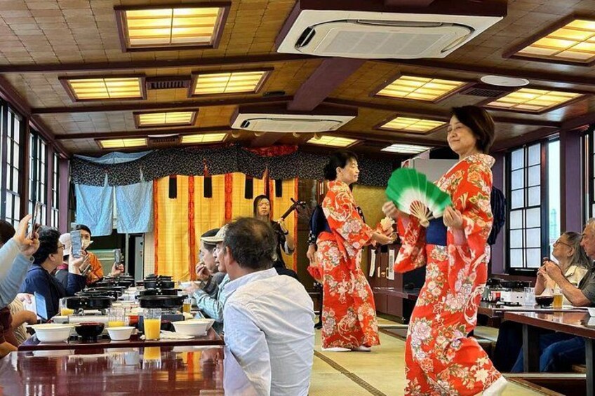 Een traditionele Japanse setting met mensen in kimono's, een houten tafel met gerechten en een podium met microfoon. 4