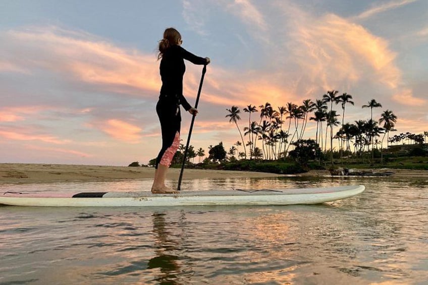 A person paddleboarding on calm water with a sunset and palm trees in the background. 4