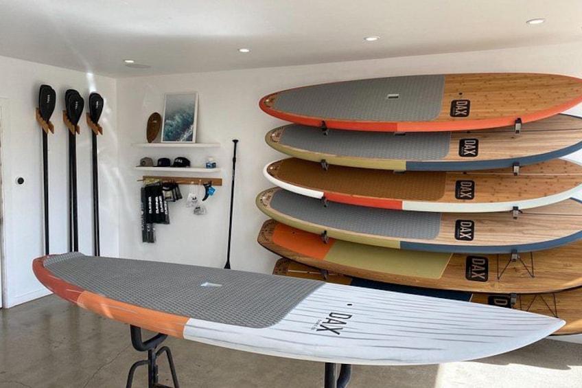 A room with several paddleboards stacked vertically and horizontally, a paddleboard in the foreground, and a wall-mounted rack with paddles. 2