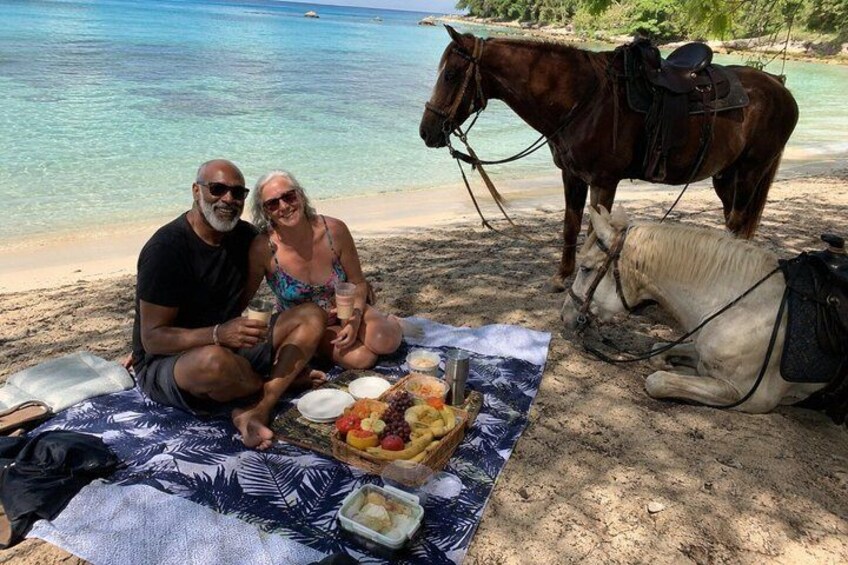 Two people sitting on a blanket with a picnic setup on a beach, a horse, and a calm sea in the background. 5