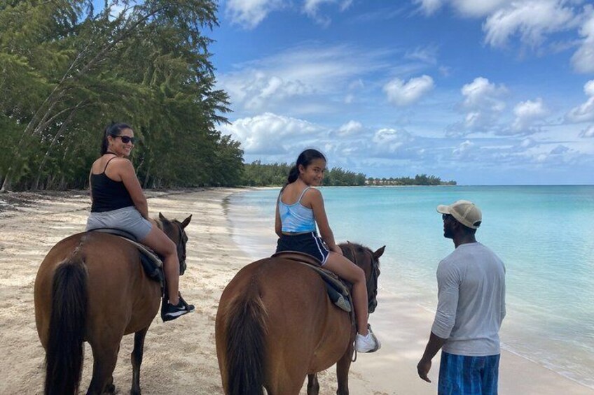 Two people riding horses on a beach with clear water and trees in the background. 3