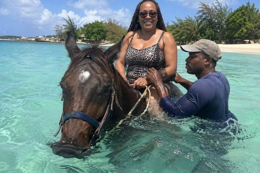 A person riding a horse in shallow turquoise water with a beach and palm trees in the background. 4