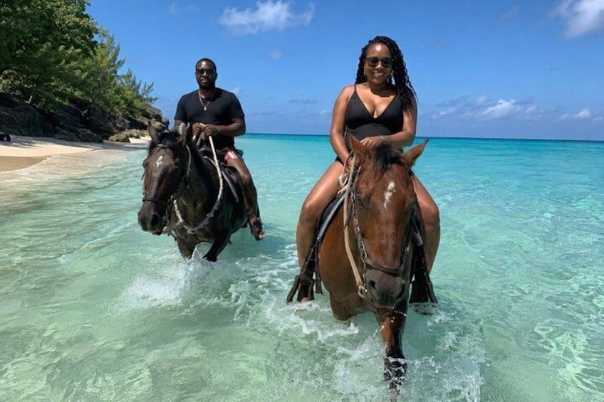 Two people riding horses through shallow, clear water near a beach with trees and a clear sky. 1