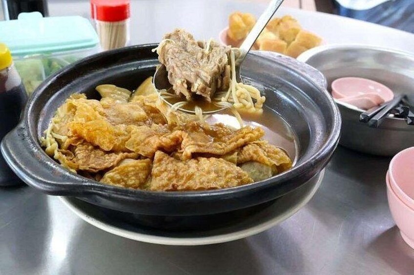 A bowl of soup with noodles and meat, a spoon lifting the meat, and a pink container in the background. 1