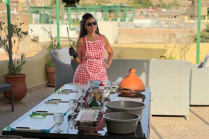Une personne portant un tablier rouge et blanc, debout près d’une table où se trouvent des ustensiles de cuisine et une casserole, sur un toit. 1