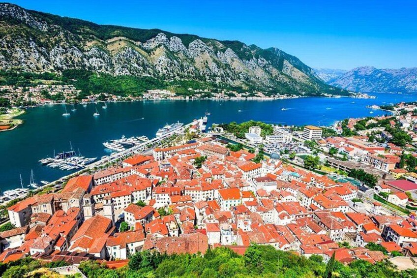 A coastal town with red-roofed buildings, a marina with boats, and mountains in the background. 2