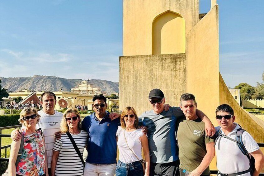 A group of people posing in front of a large yellow structure with a dome and arched entrance, with a mountainous landscape in the background. 2