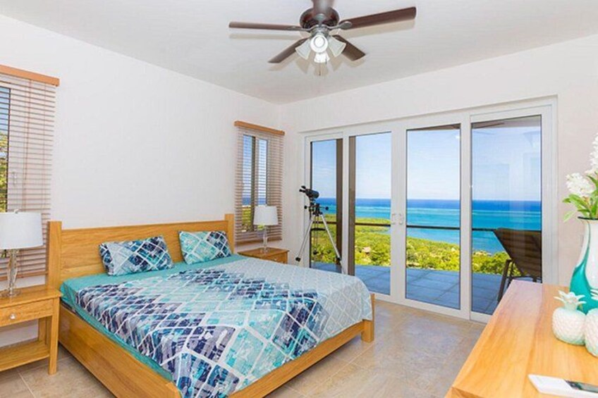 A bedroom with a bed, a ceiling fan, a nightstand, a lamp, and a view of the ocean through large sliding glass doors. 5