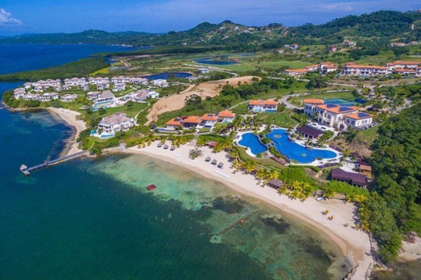 An aerial view of a coastal resort with multiple buildings, a beach, and clear blue waters. 1