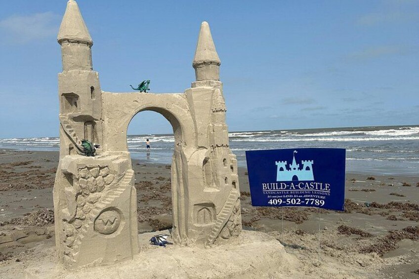 Un castillo de arena con estructura similar a un castillo, un cartel con el logotipo de un castillo y un número de contacto, y el océano al fondo. 1