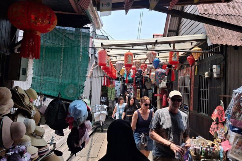 Una calle estrecha con varios puestos que venden sombreros y otros artículos, faroles rojos colgando arriba y una red verde cubriendo parte de la pared. 1