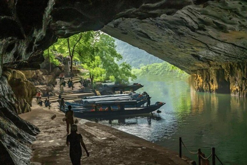 A cave entrance with boats docked on the water, a person walking, and a forested area in the background. 5