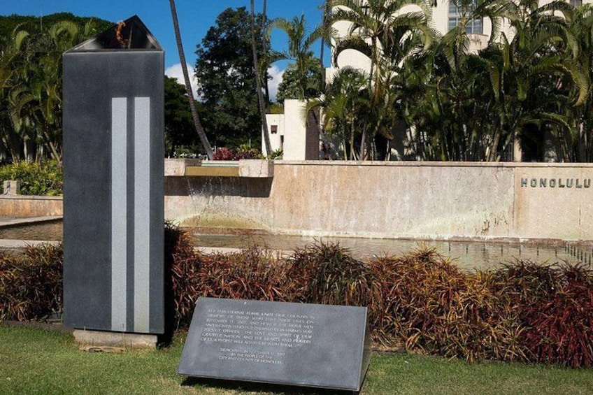 Et højt, mørkt monument med en mindeplade foran, omgivet af tropiske planter og palmer. 2