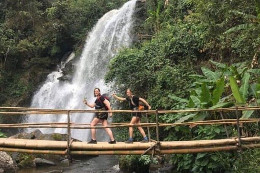 Two people on a bamboo bridge over a waterfall in a forested area. 1