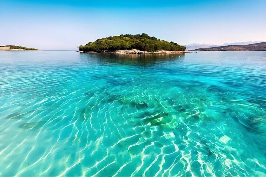 Un mar turquesa transparente, una pequeña isla con vegetación densa y verde, y montañas distantes bajo un cielo despejado. 1