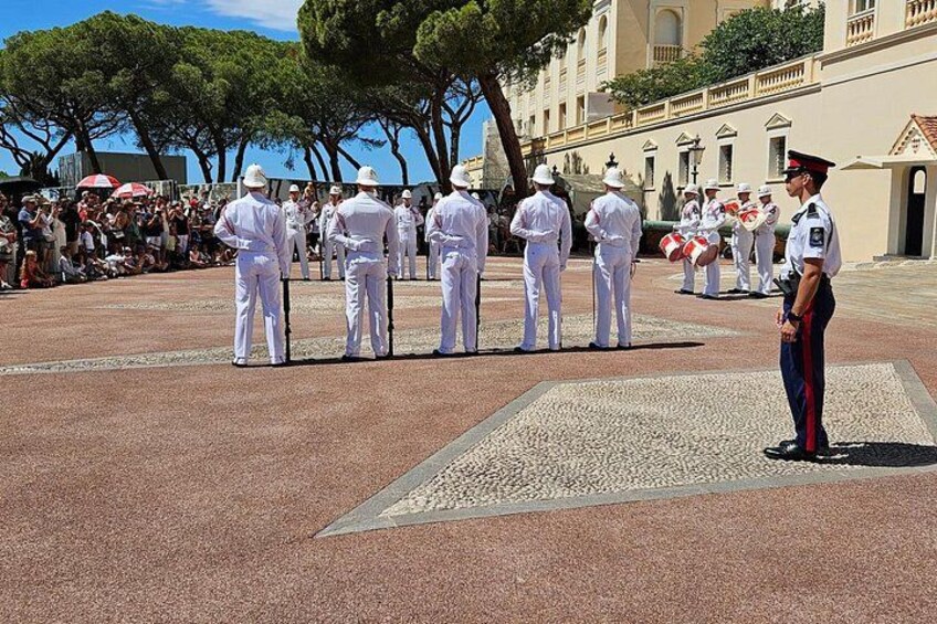 Een groep personeelsleden in uniform in een ceremoniële opstelling, staand op een versierd voetpad met bomen en een gebouw op de achtergrond. 4