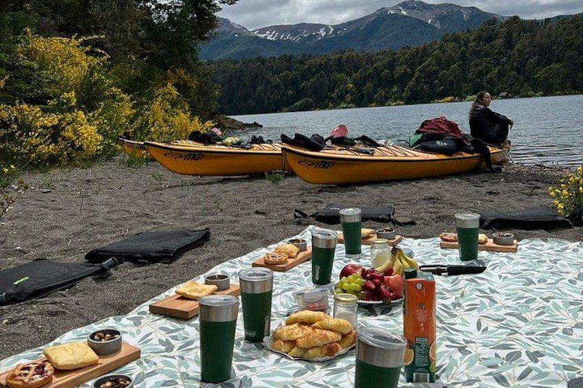 Un picnic junto al lago con una mesa dispuesta con comida y bebidas, dos canoas y una persona sentada junto al agua. 3
