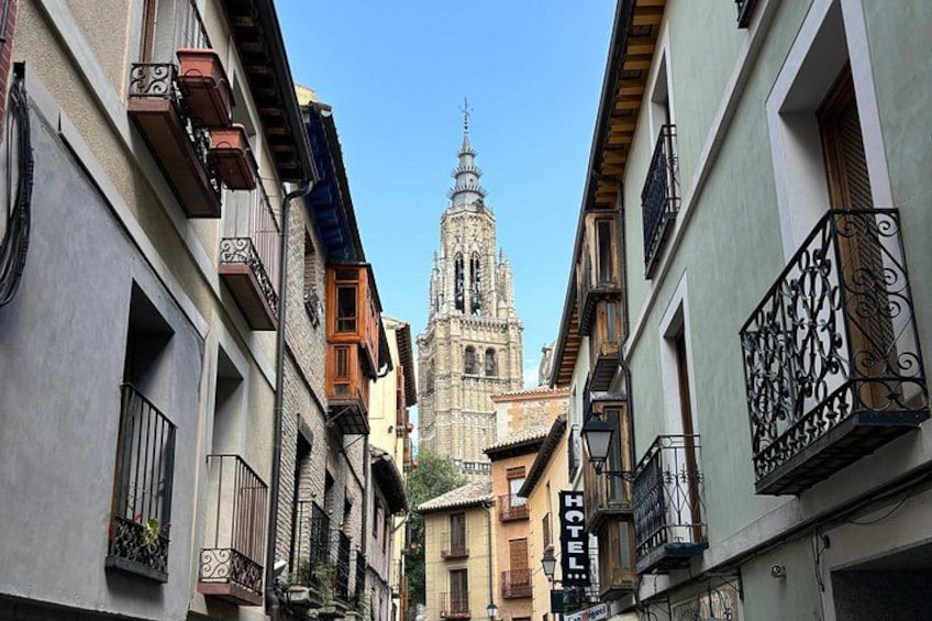 A narrow street with buildings on either side, a church with a tall bell tower, and a hotel sign. 1