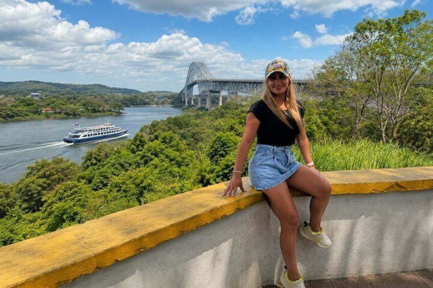 A person sitting on a concrete ledge overlooking a river with a boat and a bridge in the background. 2