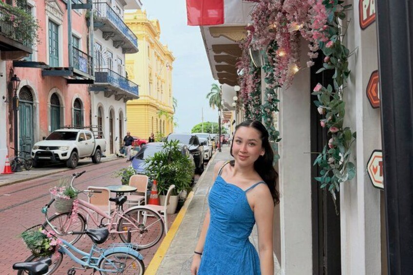 A woman in a blue dress standing next to a building with a bicycle and a cart. 4