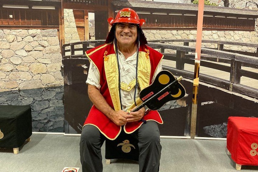 A person in a red and gold costume holding a guitar, seated on a bench with a stone wall and a wooden fence in the background. 2