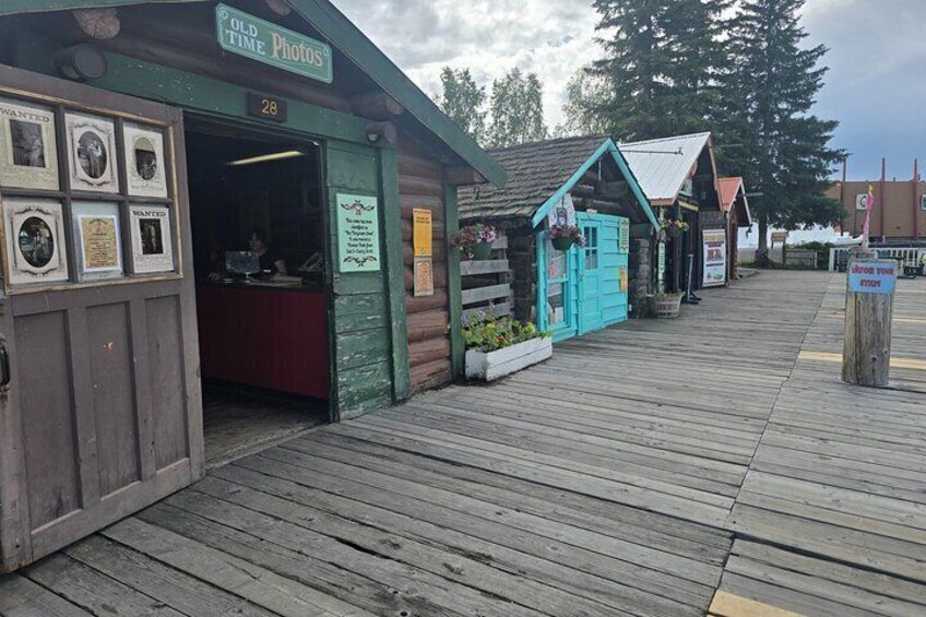 A wooden boardwalk with a green building labeled 'Old Time Photos' and a turquoise structure with plants. 4