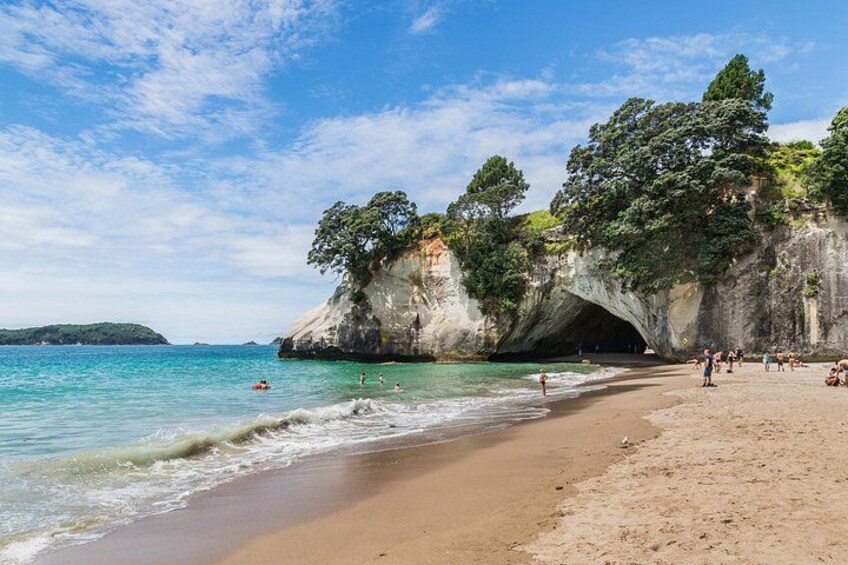 A beach with a large rock formation, clear blue water, and people swimming. 3