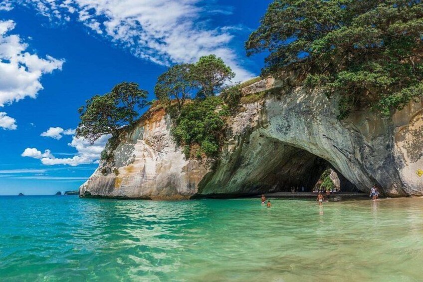 A coastal landscape with a large rock formation, clear turquoise water, and a few people swimming. 2