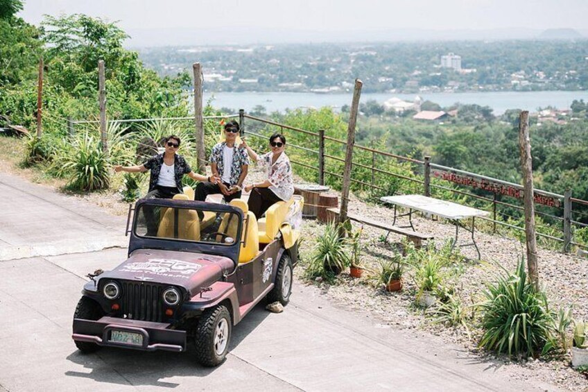 A jeep with three passengers on a paved road with a scenic view of a lake and greenery. 3
