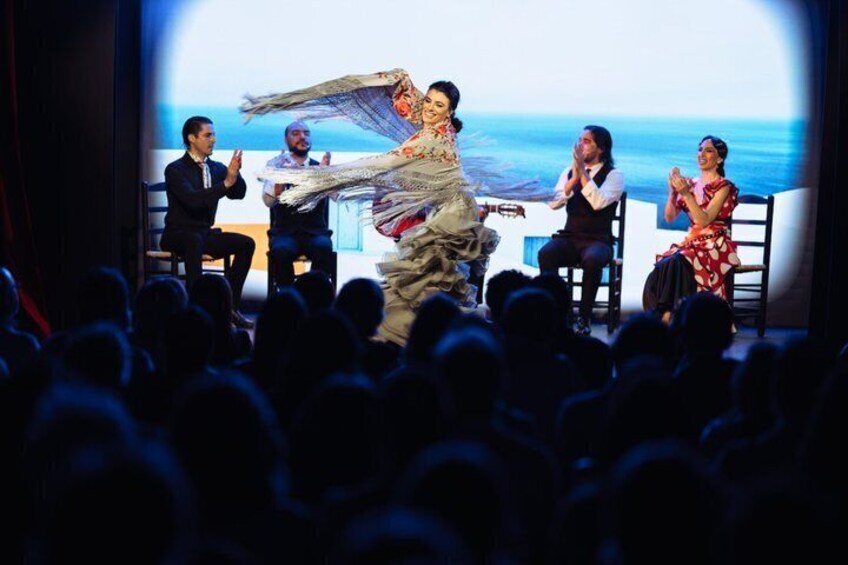 A person in a flowing garment performing on stage with an audience and panelists. 4