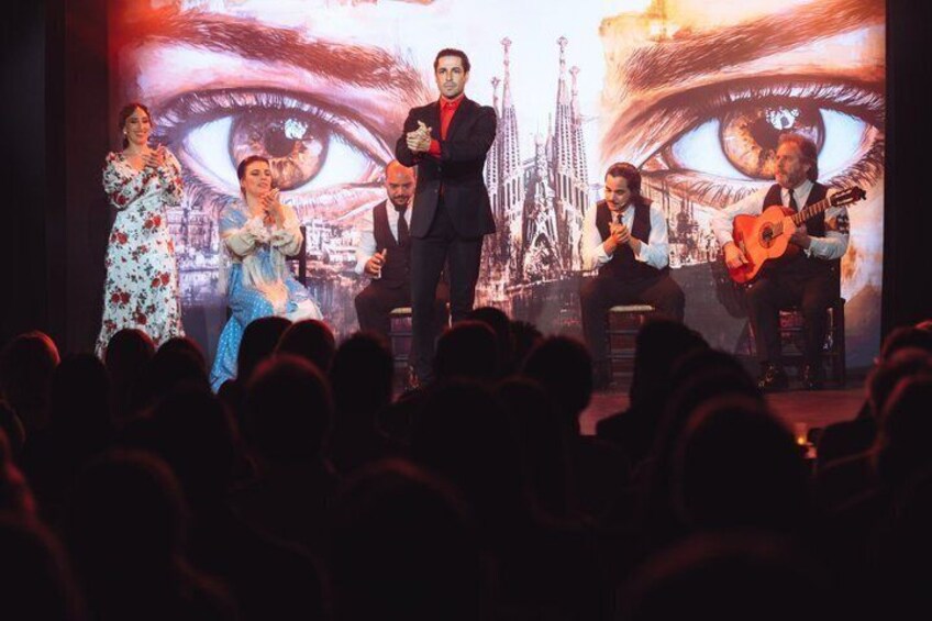 A stage performance with a large eye graphic backdrop, a man in a suit, and two guitarists. 3