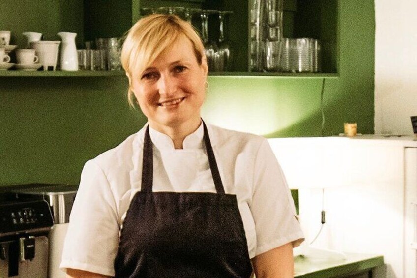 A person in a white chef's coat and black apron in a kitchen with green walls and various kitchen items in the background. 3