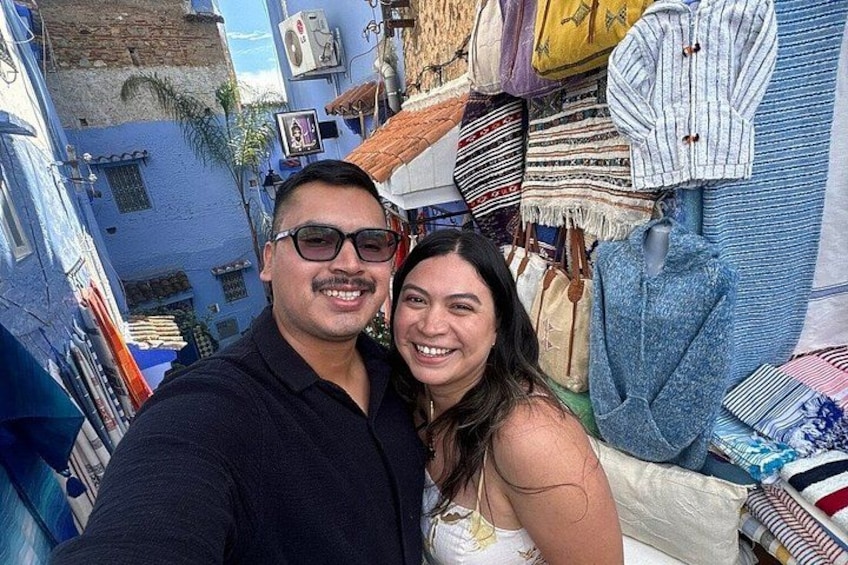 Una calle estrecha con edificios pintados de azul, un hombre y una mujer tomándose un selfie, y varios textiles exhibidos en las paredes. 3