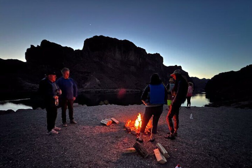 Personas alrededor de una fogata al anochecer, cerca de un lago rocoso. 1