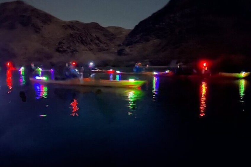 Escena nocturna con barcos reflejando luces de colores en aguas tranquilas, rodeada de montañas. 5