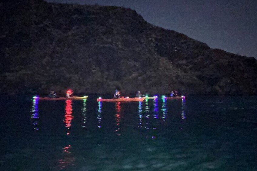 Escena nocturna con un bote iluminado por luces de colores que se reflejan en el agua, una costa rocosa y un cielo oscuro. 3