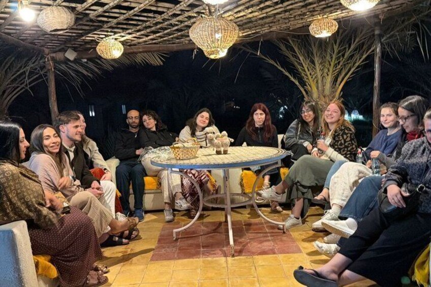 A group of people seated around a round table with a woven tablecloth, under a thatched roof structure with hanging lights. 3