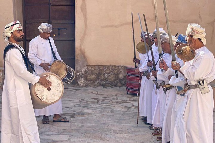 Eine Gruppe von Männern in traditioneller weißer Kleidung spielt Trommeln und hält Schwerter auf einem Innenhof. 5