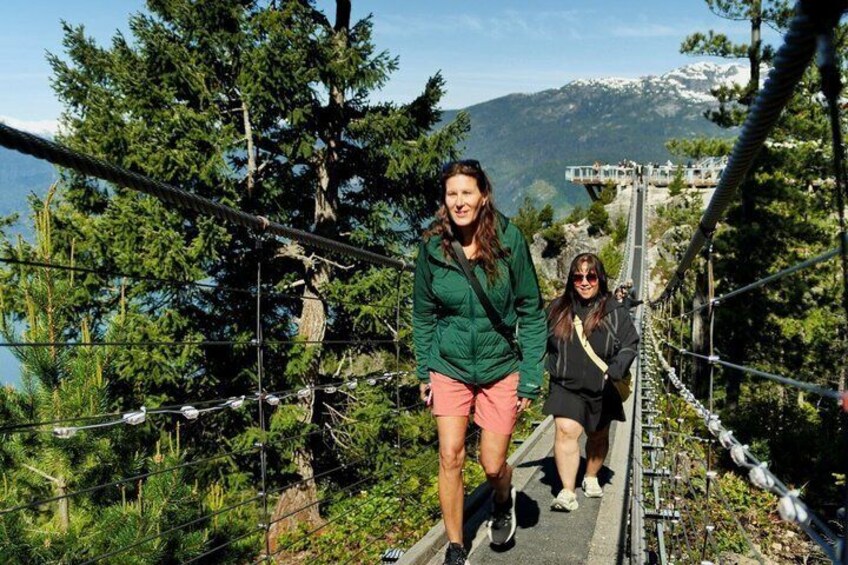 Zwei Personen gehen über eine Hängebrücke, im Hintergrund eine gebirgige Landschaft. 1